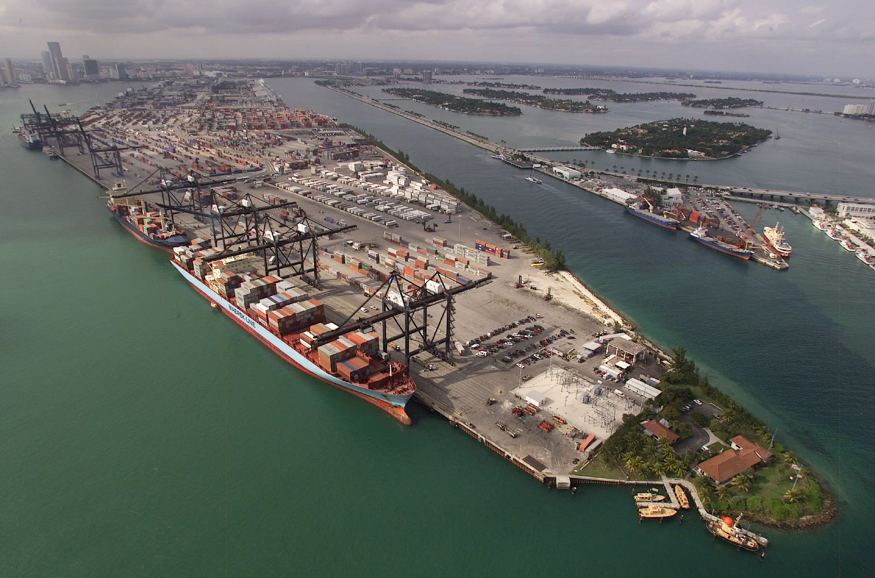 Aerial photograph of a container port: rows of stacked shipping containers beside gantry cranes and moored vessels.